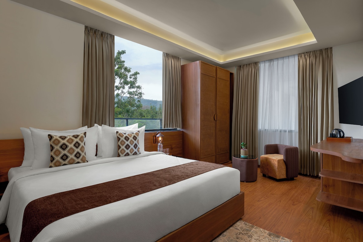 A hotel room with a large bed, a wooden wardrobe, a seating area with a chair, and a television.