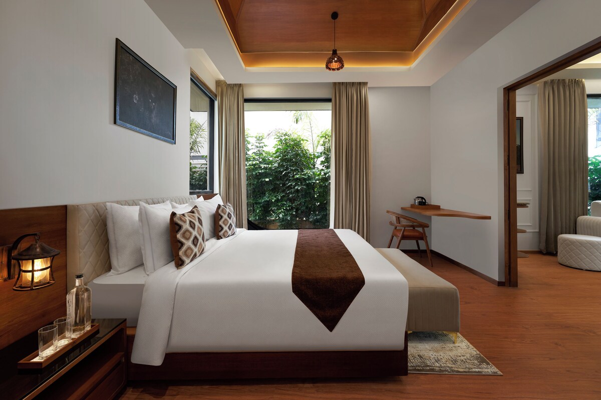 A modern hotel room with a large bed, bedside table, and a view of greenery outside.