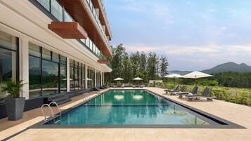 A swimming pool with lounge chairs and umbrellas, surrounded by a building with large windows and a view of mountains.