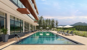 A swimming pool with lounge chairs and umbrellas, surrounded by a building with large windows and a view of mountains. - Ug The Fern Resort & Spa Chikmagalur, Series By Marriott (Chikkamagaluru)