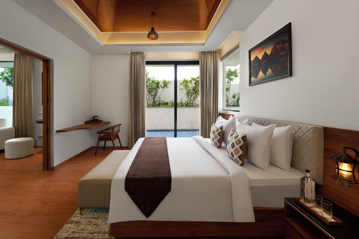 A hotel room with a large bed, a bedside table with a lamp, a view of the outdoors, and a painting on the wall.