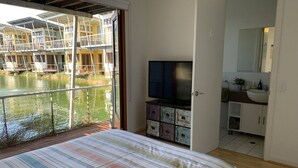 A bedroom with a bed, a television, a bathroom with a sink and mirror, and a view of a water body with buildings. - Villa #91 South Shores ~ The golden hour (Normanville)