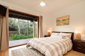 A bedroom with a large bed, two bedside tables with lamps, a painting on the wall, and a sliding glass door leading to a garden.