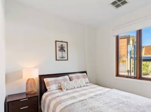 A bedroom with a bed, a nightstand, a lamp, a framed picture on the wall, and a window with a view of buildings.