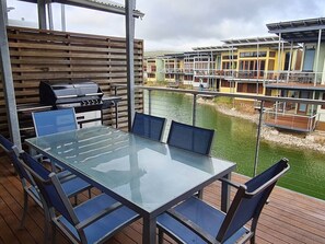 A deck with a glass-top table and chairs, a grill, and a view of a water body and buildings in the background.