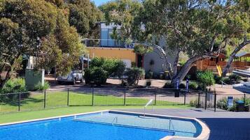 A swimming pool with a curved edge, surrounded by a black fence, trees, and a building in the background.