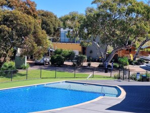 A swimming pool with a curved edge, surrounded by a black fence, trees, and a building in the background.
