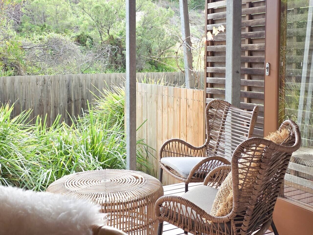 A wicker outdoor seating area with a round table, a wooden fence, and a view of greenery.