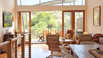 A spacious room with a large sliding glass door leading to a balcony, wooden furniture, and a view of greenery.
