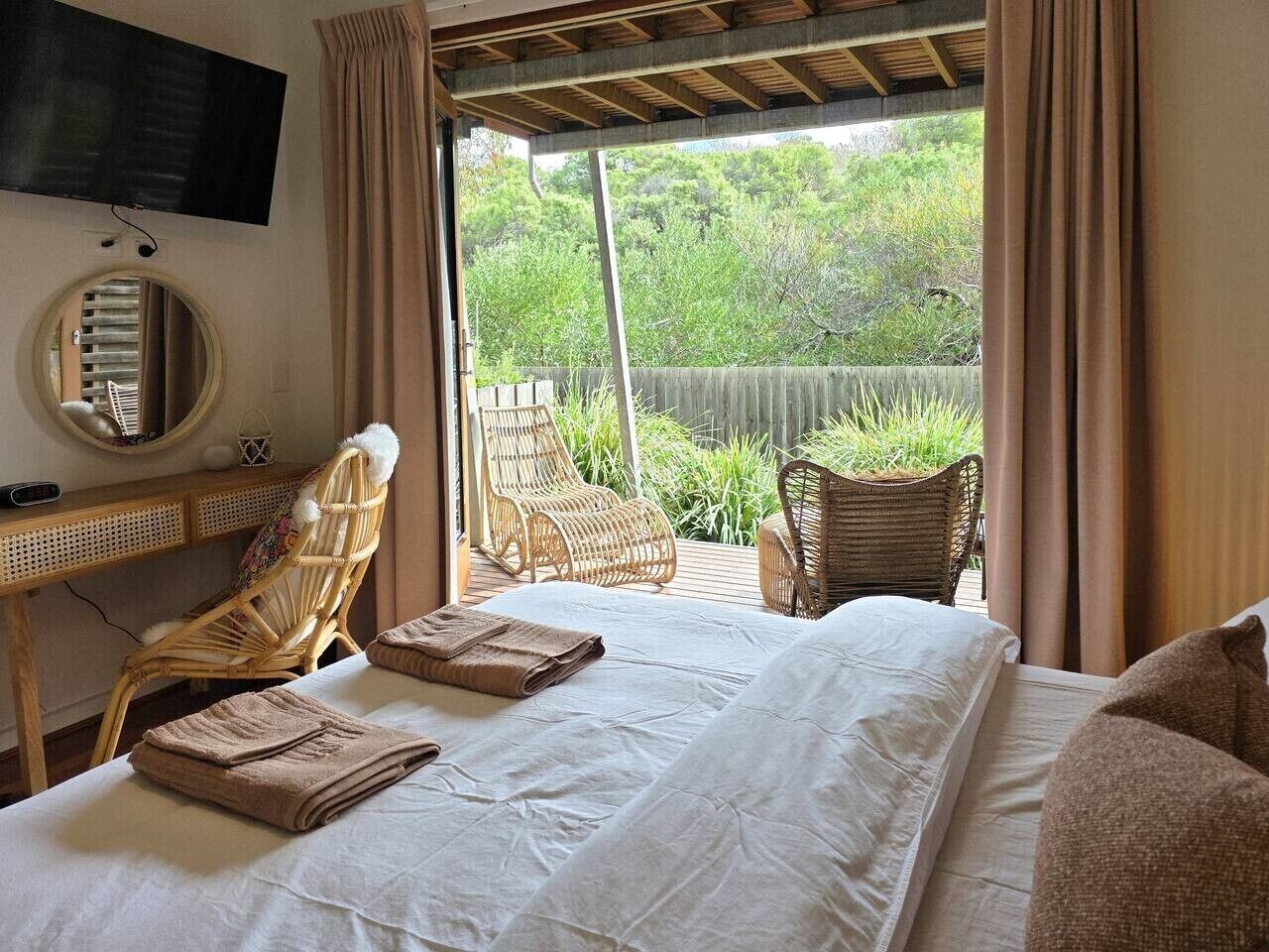 A bedroom with a bed, two wicker chairs, a desk, a TV, and a view of greenery outside.