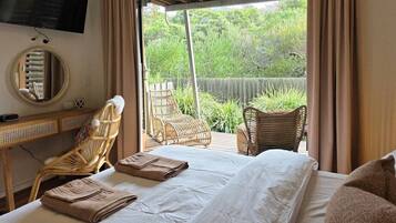 A bedroom with a bed, two wicker chairs, a desk, a TV, and a view of greenery outside.