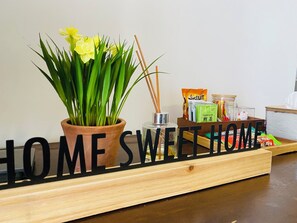 A wooden sign with 'HOME SWEET HOME' text, a potted plant with yellow flowers, a reed diffuser, and a box of tea. - Modern 2BR Condo in Bukit Bintang with Pool, Gym, Netflix, Walk Everywhere (Kuala Lumpur)