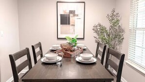 A dining room with a dark wooden table, white chairs, a framed picture on the wall, and a potted plant. - Central Arlington Gem Near AT&T Stadium & Texas Live 2BR (Arlington)