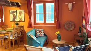 Un intérieur rustique avec des poutres apparentes, un coin repas comprenant une table et des chaises, un canapé et une fenêtre avec des volets.