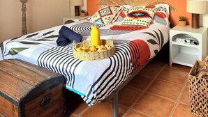 Un lit avec une tête de lit ornée d’un motif de feuilles, une table de chevet avec une lampe, un tableau accroché au mur et un panier en osier.