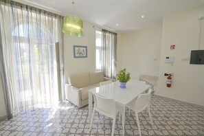 Ein Zimmer mit einem weißen Tisch, einem beigefarbenen Sofa, einer Topfpflanze, einem Feuerlöscher und einem fußbodenähnlichen Dekor.