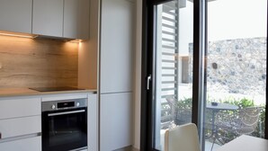 A modern kitchen with built-in appliances, a dining area with a table and chairs, and a view of a stone wall and outdoor seating. - Villa Elegance in Nikiti (Sithonia)