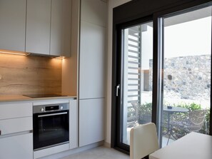 A modern kitchen with built-in appliances, a dining area with a table and chairs, and a view of a stone wall and outdoor seating. - Villa Elegance in Nikiti (Sithonia)