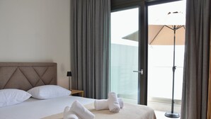 A hotel room with a bed, pillows, a bedside table with a lamp, and a view through a glass door to an outdoor area with a sun umbrella. - Villa Elegance in Nikiti (Sithonia)