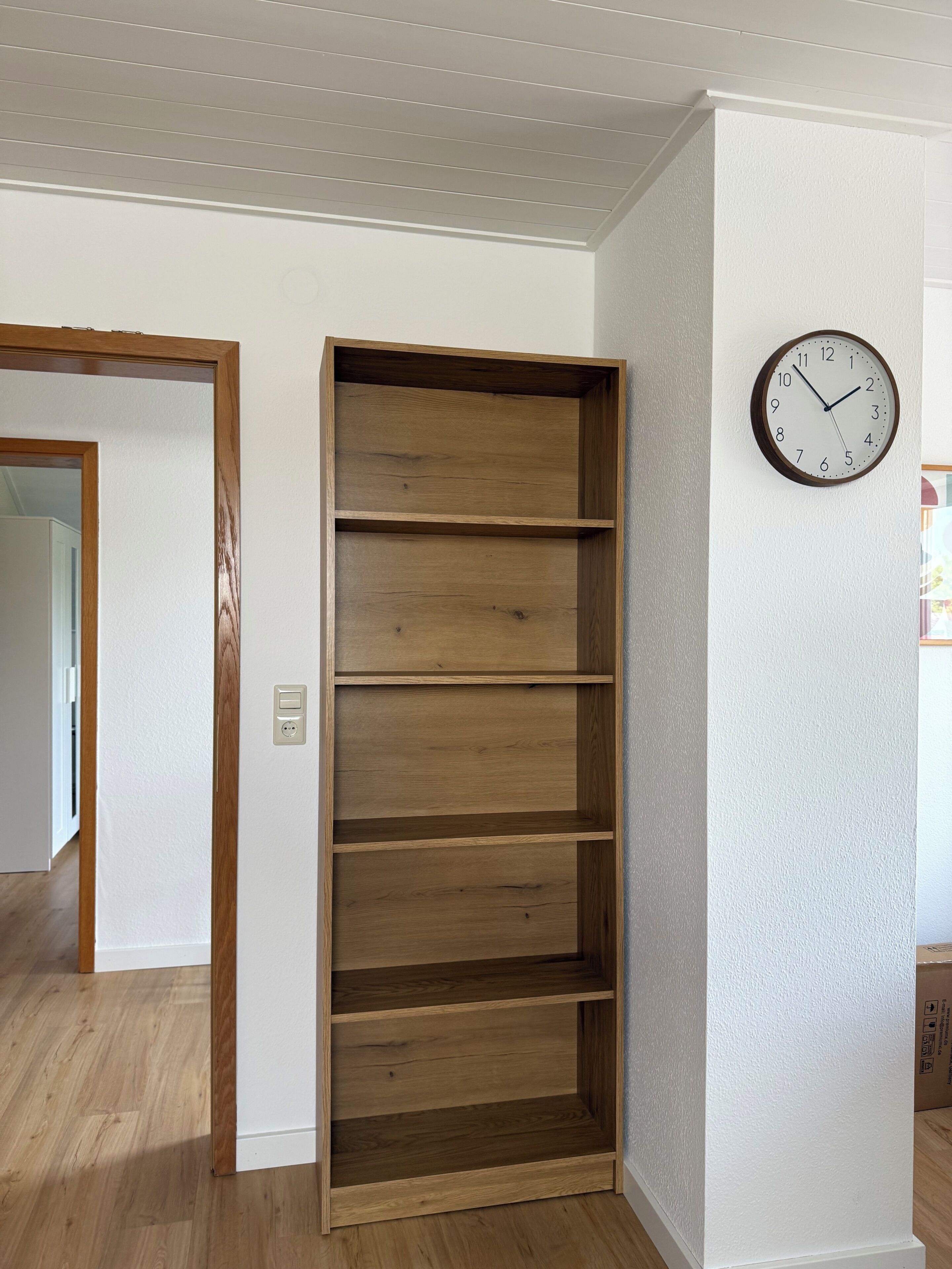 A room with a wooden shelving unit, a wall clock, and a door leading to another room.