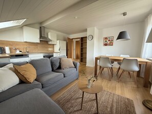 Ein modernes Wohnzimmer mit grauem Sofa, einem Esstisch mit Stühlen, einer Küchenzeile mit Schränken und einem Herd sowie einem großen Fenster.