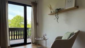 Una stanza con una porta scorrevole in vetro che dà su un balcone, un tavolino rotondo con un vaso, una sedia e uno scaffale con una pianta e una cornice.