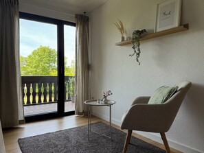 Ein Zimmer mit einer Schiebetür zum Balkon, einem kleinen runden Tisch mit einer Vase, einem Stuhl und einem Regal mit Pflanze und gerahmtem Bild.