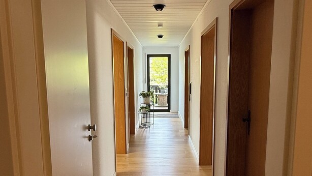 Un corridoio con pavimento in legno e pareti bianche, che conduce a una porta da cui si vede un albero.