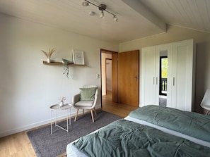 Ein Schlafzimmer mit Bett, Sessel, kleinem Tisch, Pflanzenregal, Tür und Kleiderschrank.