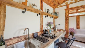 A kitchen with a stainless steel sink, wooden shelves, a kettle, and a dining area with a chair and a sofa. - 1 bedroom accommodation in Corwen (Corwen)