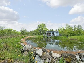 Een huis op palen boven een vijver, met een terras en stoelen, omringd door groen en bomen.