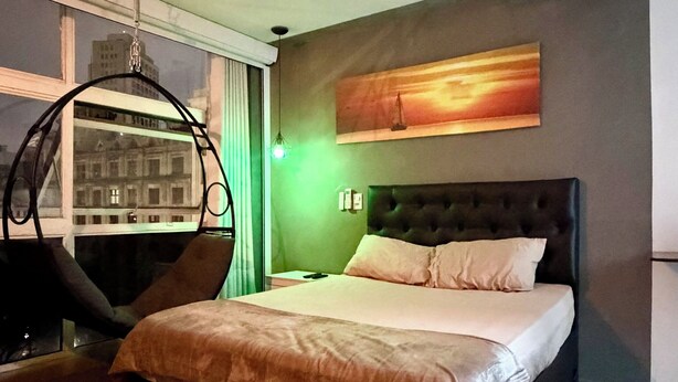 Quarto de hotel moderno com uma cama grande, uma cadeira suspensa, vista da cidade pela janela e um quadro na parede.