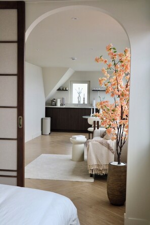 Un interior moderno con suelo de madera, un sofá blanco, una planta grande en maceta y una cocina al fondo.