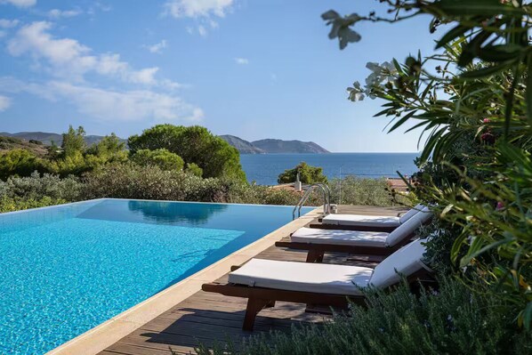 A clear blue pool with wooden deck and lounge chairs, surrounded by greenery and overlooking the sea. - Villa Valli Athenian Riviera, (Anavissos)