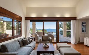 A spacious living room with a large sofa, a coffee table, and a view of the sea through large sliding glass doors. - Villa Valli Athenian Riviera, (Anavissos)