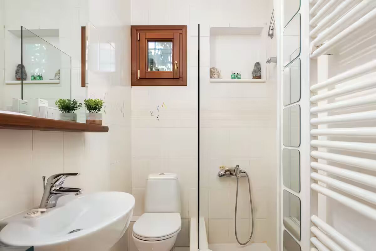 Une salle de bain avec un lavabo, des toilettes et une douche, le tout de couleur blanche. On y trouve une petite fenêtre avec un cadre en bois, ainsi qu’une étagère contenant des articles de toilette.