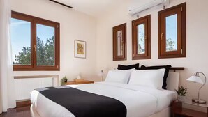 A bedroom with a large bed, wooden headboard, multiple windows, a radiator, a bedside table with a lamp, and a framed picture on the wall. - Villa Valli Athenian Riviera, (Anavissos)