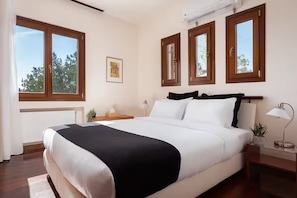 A bedroom with a large bed, wooden headboard, multiple windows, a radiator, a bedside table with a lamp, and a framed picture on the wall. - Villa Valli Athenian Riviera, (Anavissos)