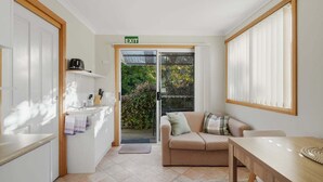 Een kamer met een kitchenette, een bank, een eettafel en een glazen schuifdeur die uitkomt op een tuin.