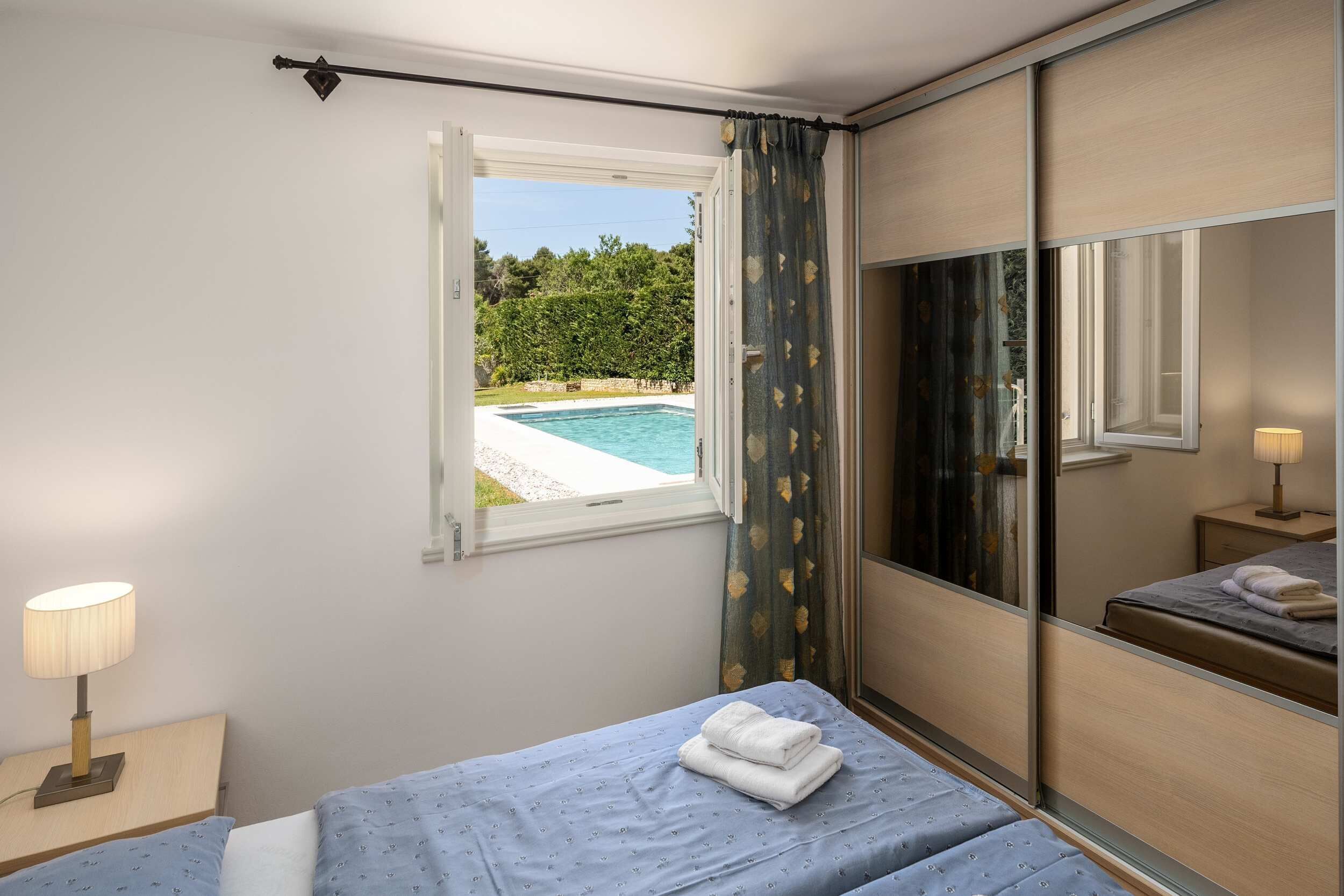 Une chambre à coucher avec un lit, une table de chevet, une lampe, une fenêtre avec des rideaux et une vue sur une piscine.