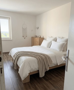 A bedroom with a bed, wooden floor, a bedside table with a lamp, a wall-mounted dreamcatcher, and a window with blinds. - Stylish 2-Bedroom Crikvenica Apartment with AC and Coastal Charm (Crikvenica)
