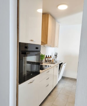 A compact kitchen with a black oven, white cabinets, a wooden upper cabinet, and a countertop with kitchen utensils. - Stylish 2-Bedroom Crikvenica Apartment with AC and Coastal Charm (Crikvenica)