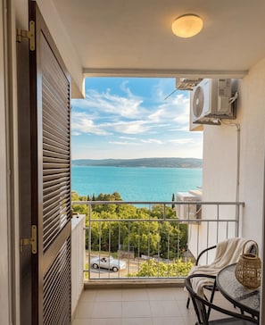A balcony with a view of a body of water and distant hills, a table with a chair, and an air conditioning unit. - Stylish 2-Bedroom Crikvenica Apartment with AC and Coastal Charm (Crikvenica)