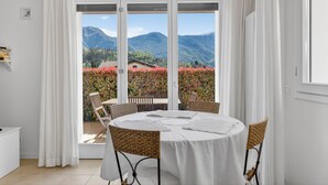 A round table with white tablecloth and wicker chairs, a white cabinet, and a view of mountains and red flowers through large windows. - Apartment with pool, parking space, 2 bedrooms, pedestrian area (Lenno)