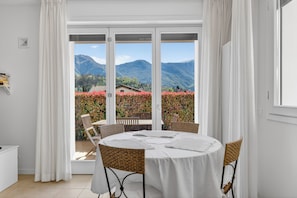 Ein runder Tisch mit weißer Tischdecke und Korbstühlen, ein weißes Sideboard und ein Blick auf Berge und rote Blumen durch große Fenster.