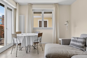 Ein Zimmer mit Esstisch, Stühlen, einer Couch, einem Fenster mit Vorhängen und einer Heizung.