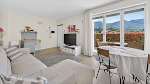 A modern living room with a sofa, TV, and dining table, large windows with a mountain view. - Apartment with pool, parking space, 2 bedrooms, pedestrian area (Lenno)