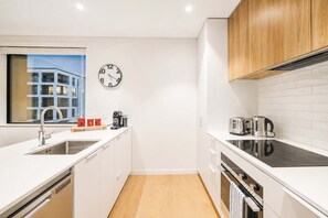 A modern kitchen with white cabinetry, a stainless steel sink, a built-in oven, and a window with a view of a building. - Central City Stay | 2BR + Balcony | Parking (Christchurch)