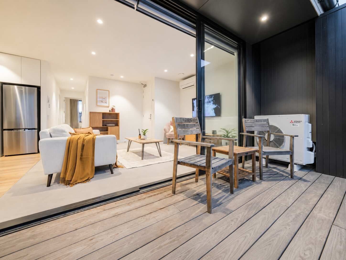 A modern living room with a sofa, coffee table, and chairs, a kitchen with a refrigerator, and a large window.