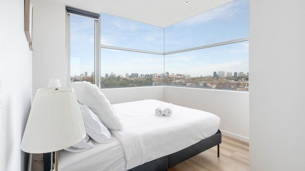 Un dormitorio moderno con vista a la ciudad, una cama con ropa de cama blanca, una lámpara de noche y un paisaje urbano visible a través de grandes ventanales.